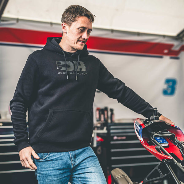 Man wearing a black hoodie with a logo, standing next to a motorcycle in a garage.