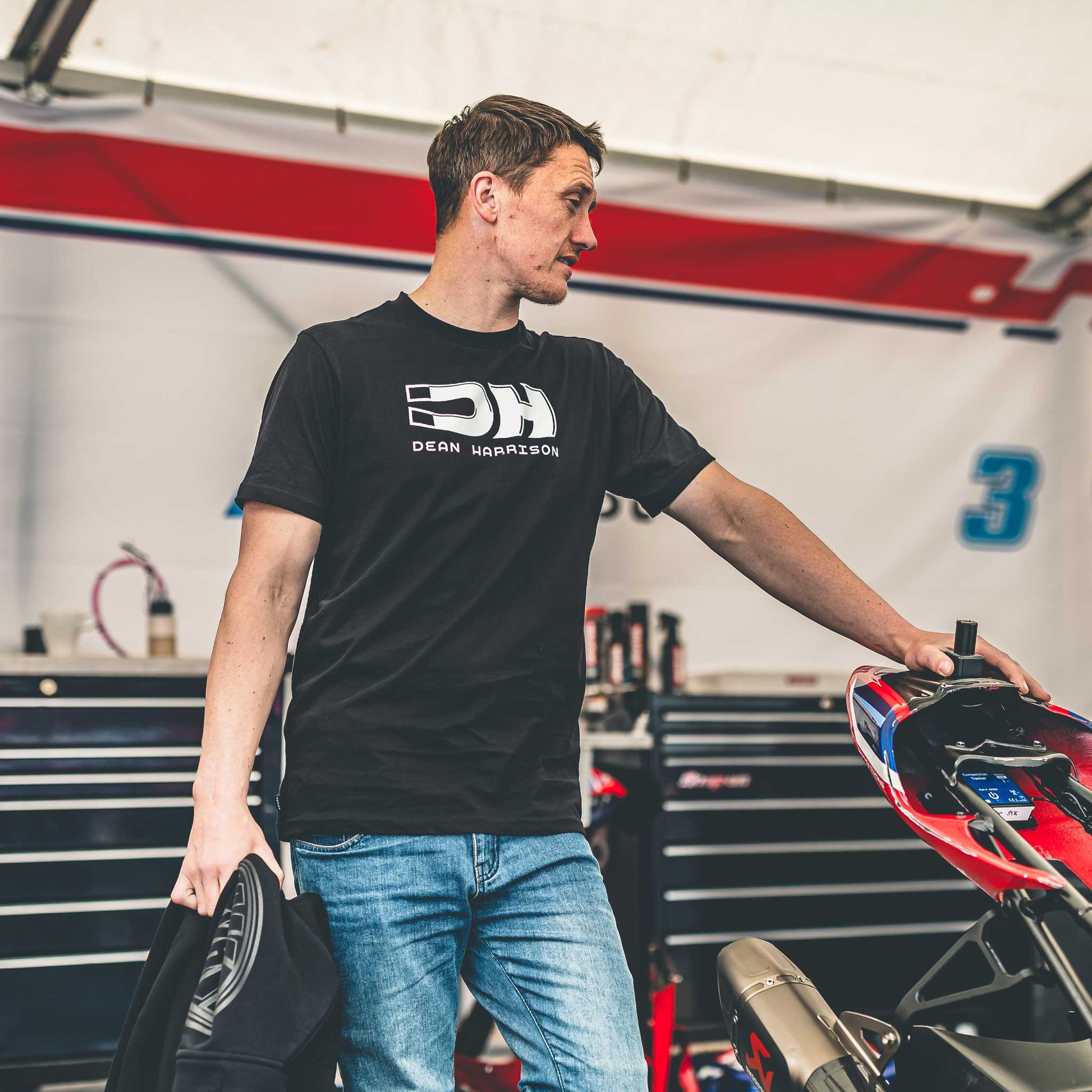 Man wearing a black t-shirt with a logo, standing in a garage with tools and equipment.