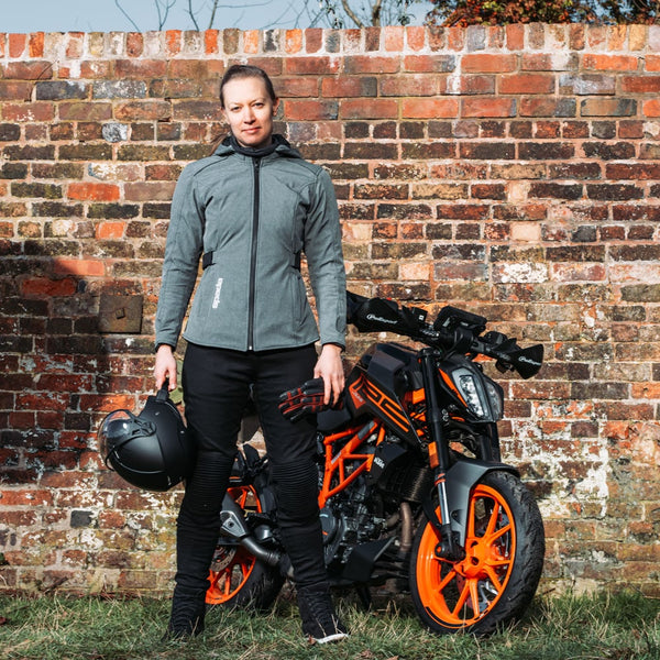 Woman wearing grey ladies motorbike jacket holding helmet next to black and orange motorcycle, styled with casual motorbike trainers