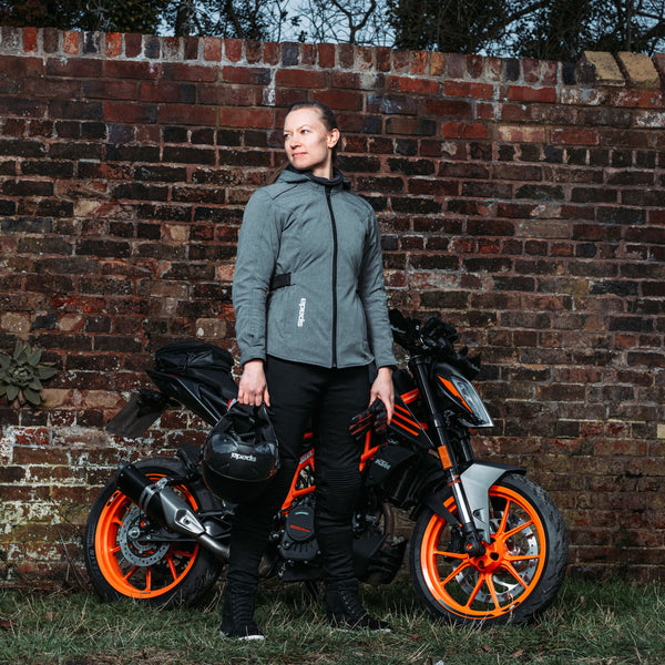 Side angle of woman in grey womens motorcycle jacket holding helmet beside black and orange motorbike, showcasing ergonomic fit and style