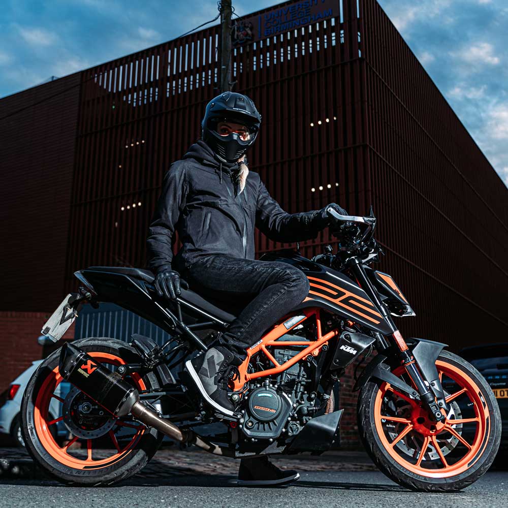 Woman rider sitting on black and orange motorcycle wearing unisex black motorbike jacket, breathable, waterproof gear for female bikers