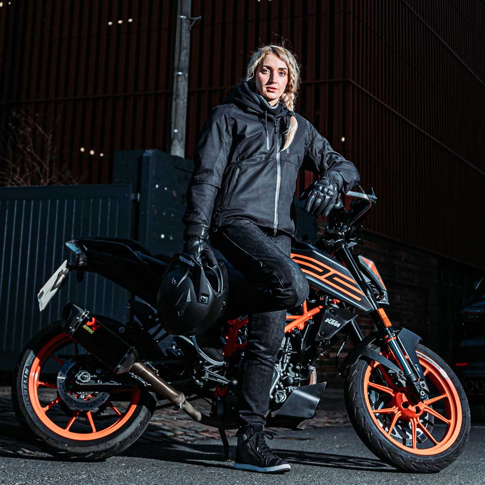 Female biker wearing Joe Matt black motorcycle jacket next to orange and black motorbike, unisex weatherproof riding gear with helmet in hand