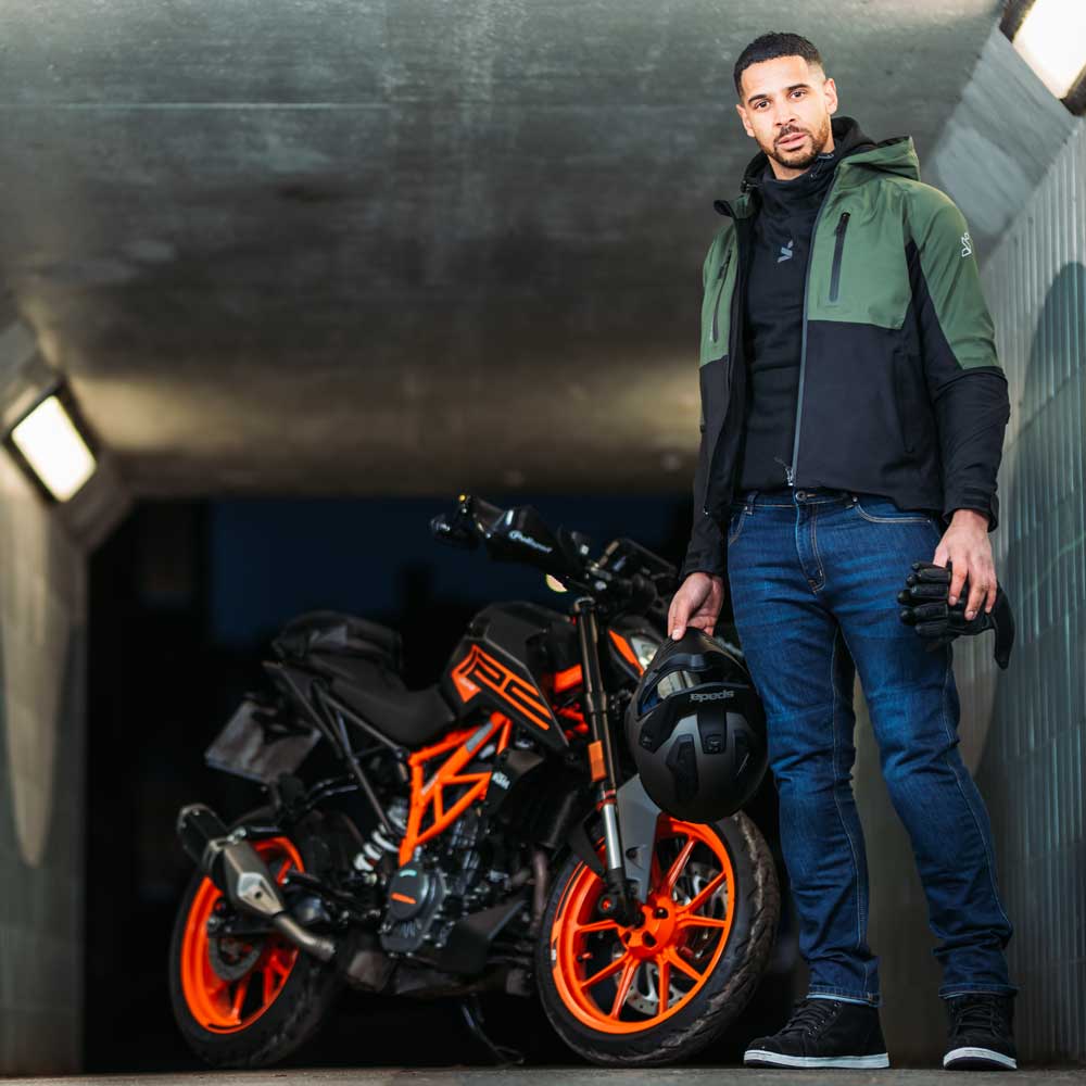 Front view of male biker wearing unzipped green and black motorbike jacket, styled with blue riding jeans and trainers next to black and orange bike