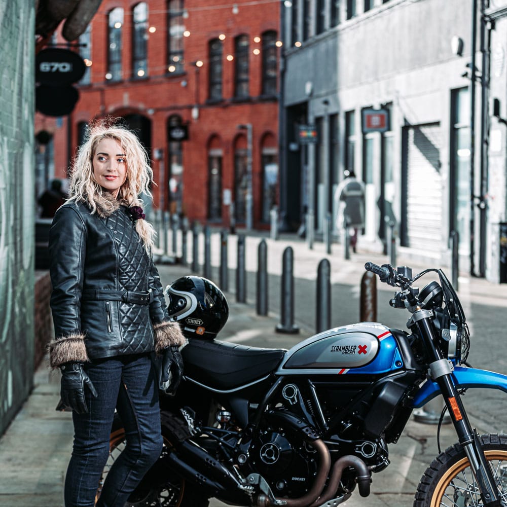 female biker wearing black leather women's motorcycle jacket with riding jeans next to a motorbike