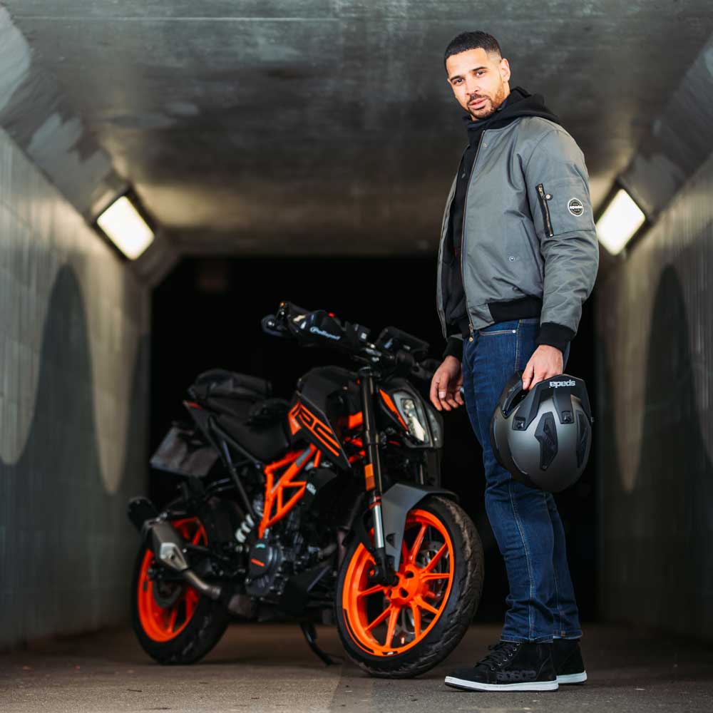 biker standing next to motorbike holding black motorcycle helmet and wearing grey bomber jacket