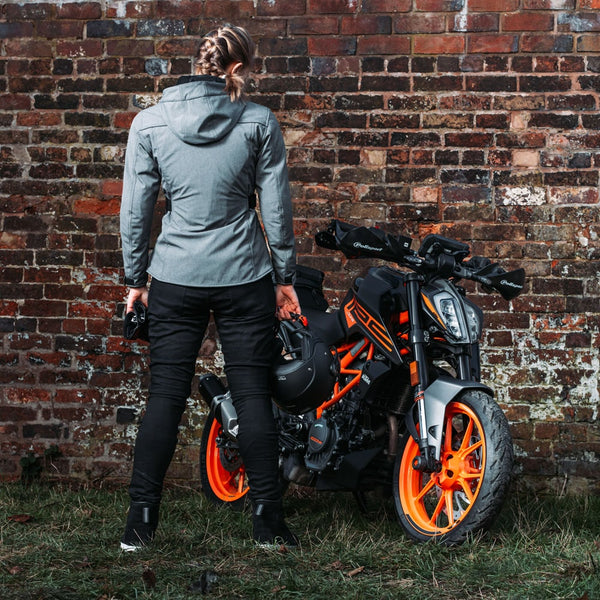 Back view of woman wearing grey ladies waterproof motorbike jacket standing next to black and orange motorcycle, highlighting adjustable waist and hip fit